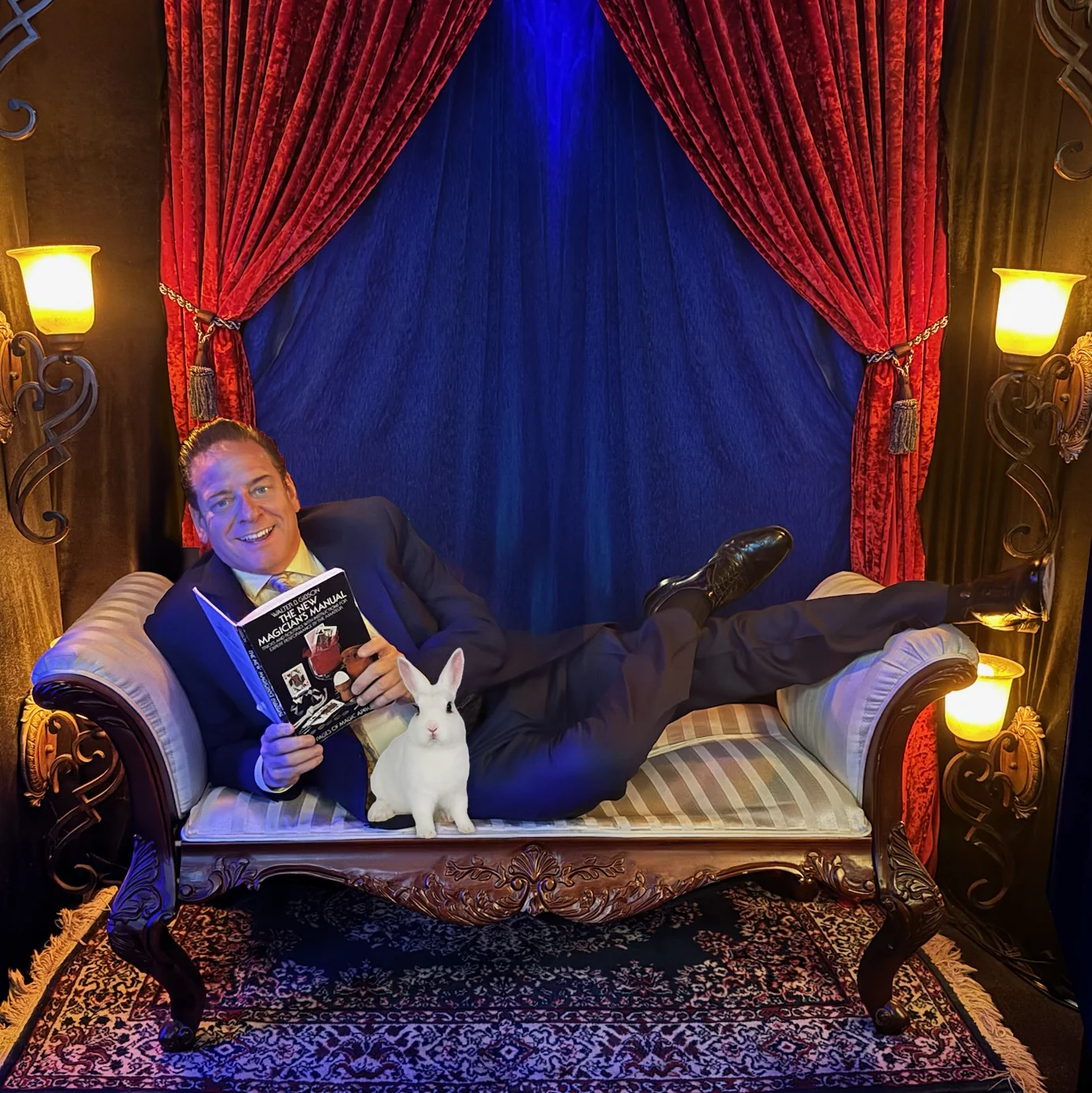 Mike Thornton relaxing with a magic book and a white rabbit against red theater curtains